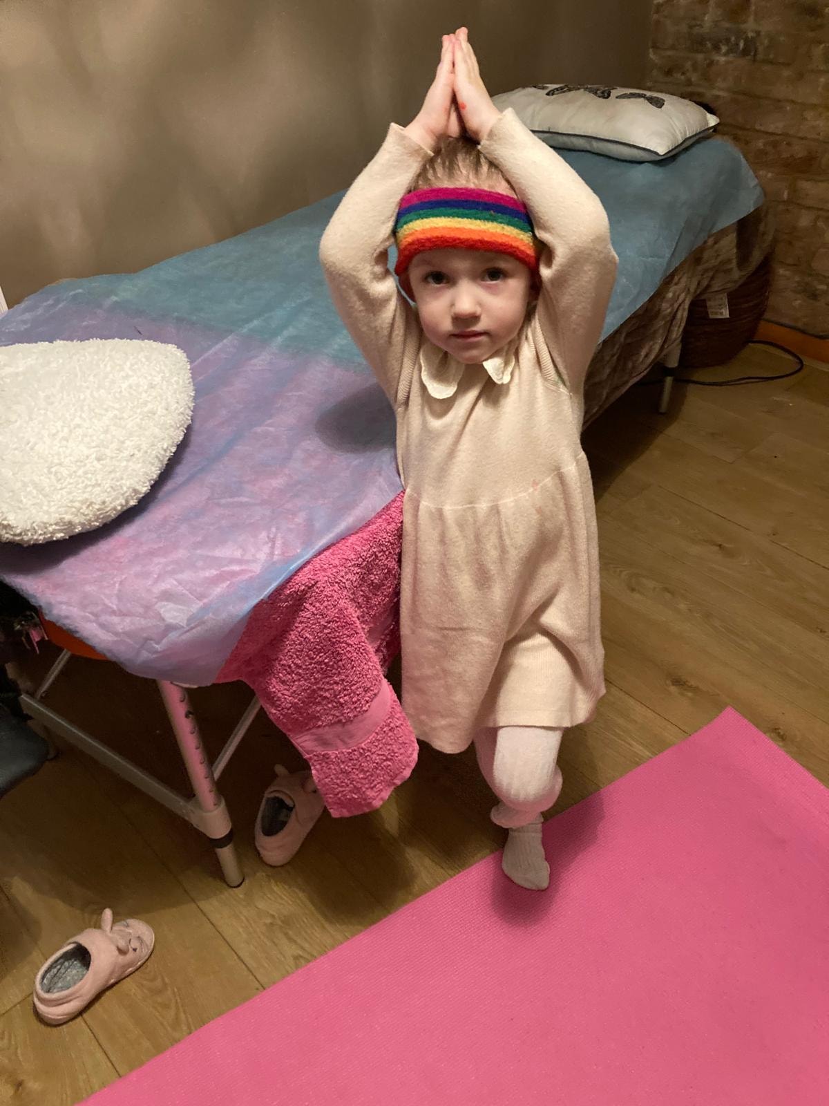 A young child in a rainbow headband practising yoga on a pink mat in the treatment room