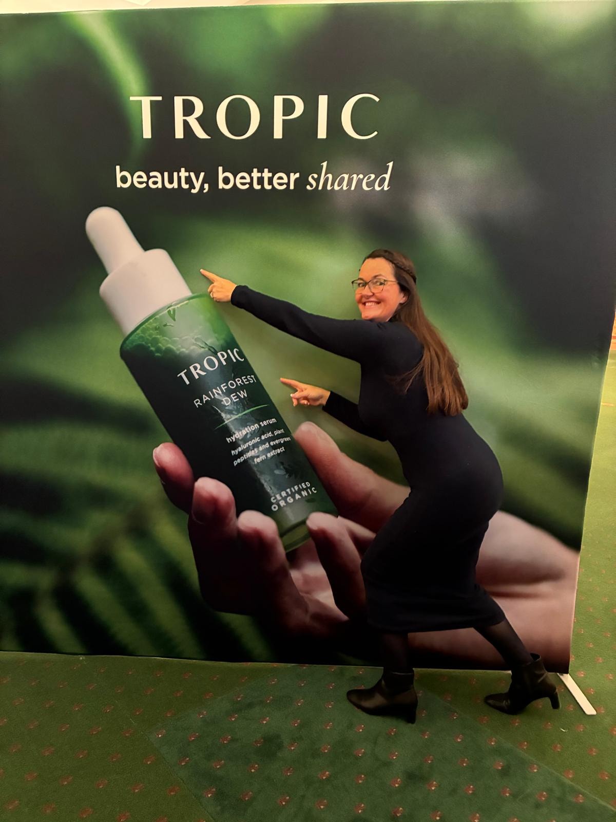 Mandie smiling and pointing at a giant Tropic Rainforest Dew serum display — beauty, better shared
